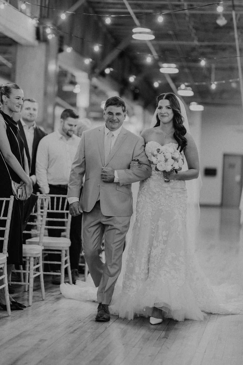 Black and white photo of bride and father walking down wedding aisle with string lights above.