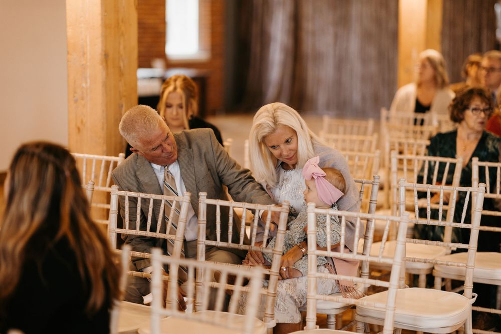 Wedding guests in white Chiavari chairs review programs during an indoor ceremony.