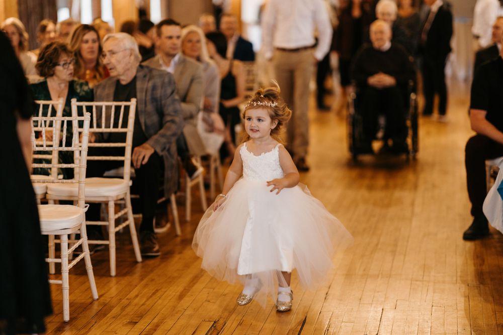 An adorable flower girl in a white tulle dress walks down the aisle at a wedding ceremony.