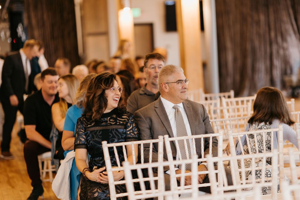 Wedding guests laughing and smiling while seated in white chairs at indoor ceremony venue.