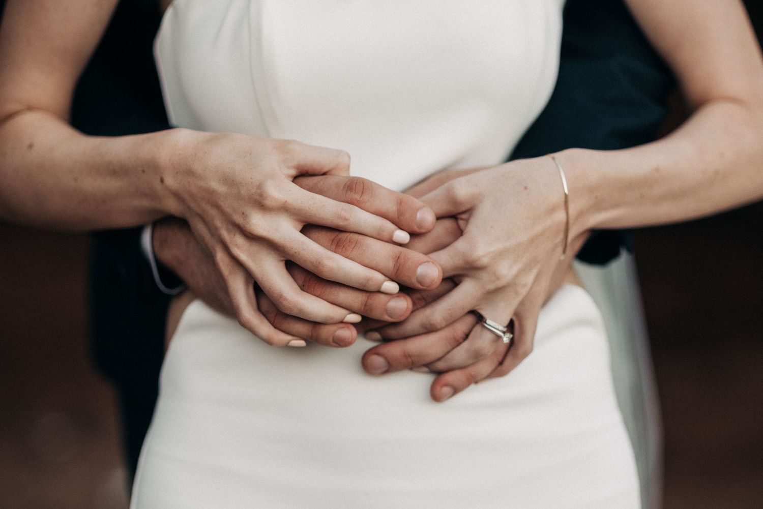 Close-up of hands with wedding rings intertwined against a white dress.