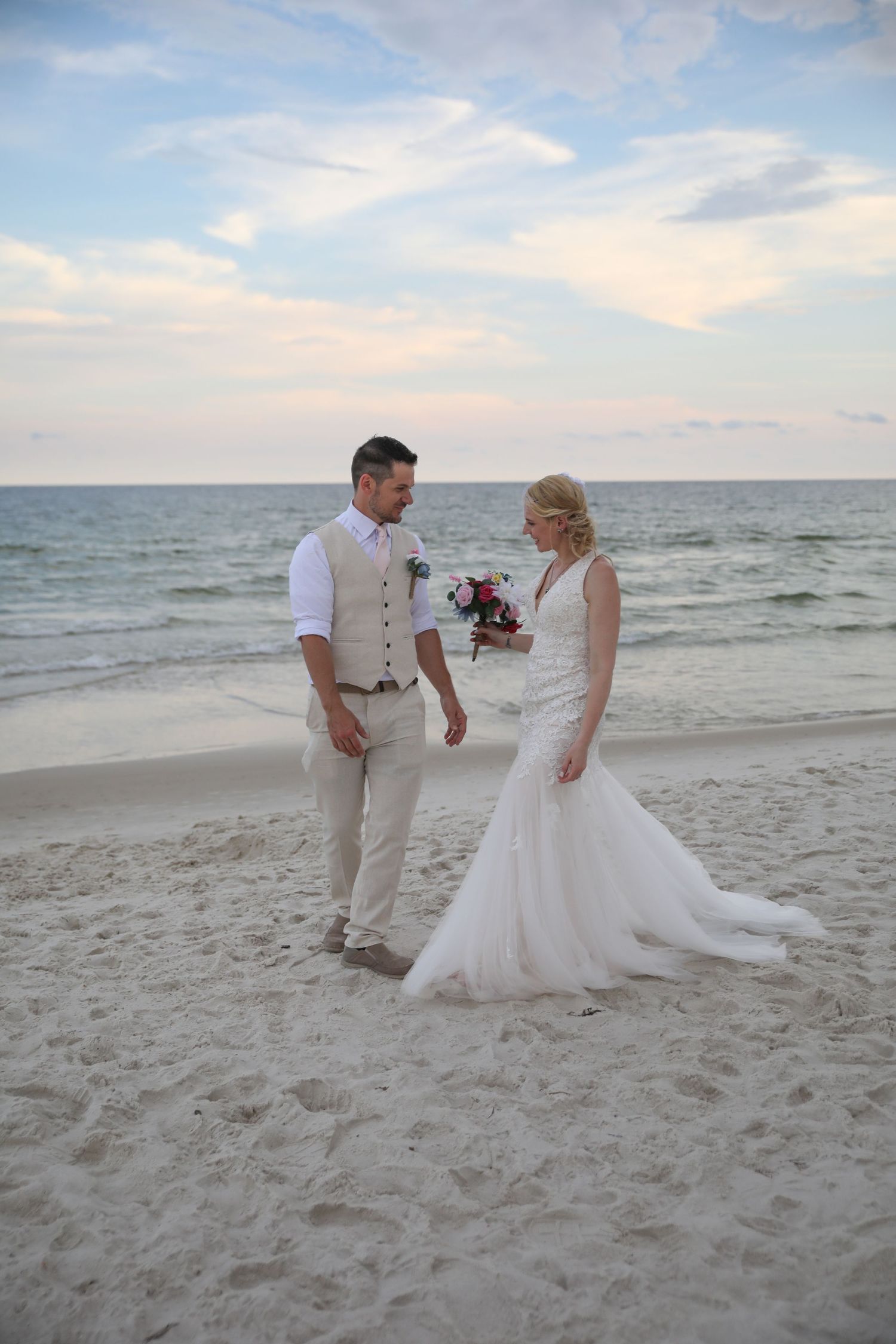A couple in light-colored wedding attire standing on a beach at dusk with waves in background.