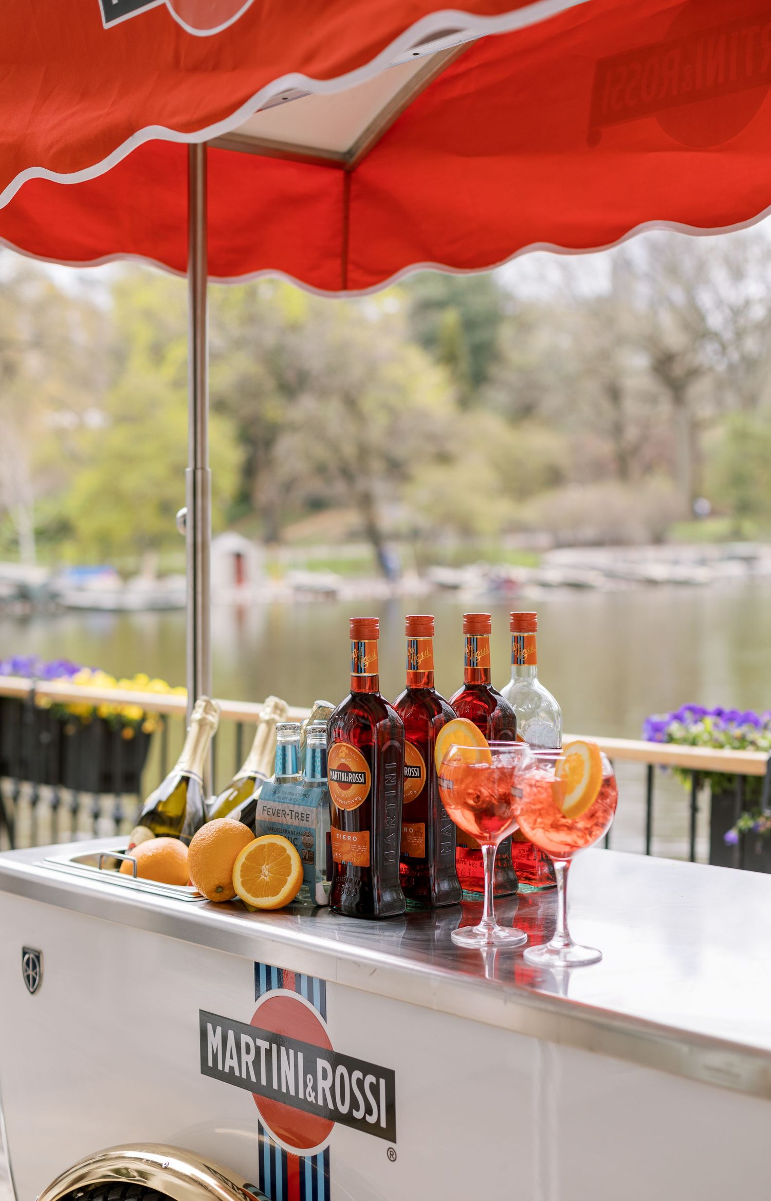 Martini & Rossi bar cart with cocktail supplies under a red umbrella on an outdoor terrace.