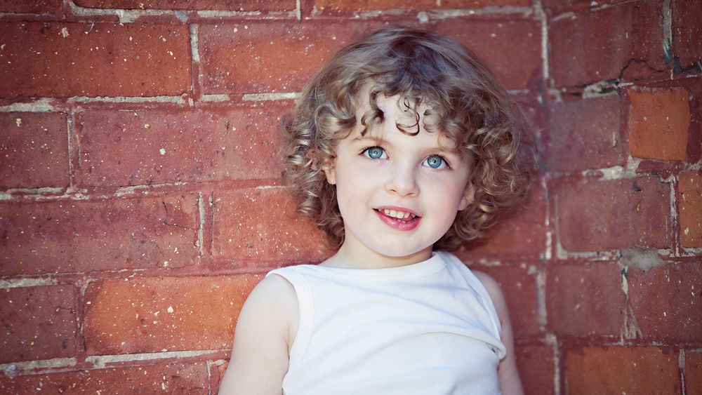 A child with curly hair smiles while leaning against a red brick wall.