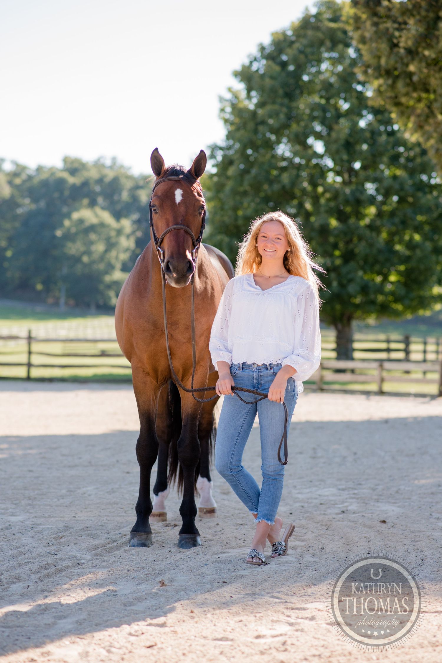 Bay horse and person in casual attire standing together in a sandy arena with fenced pasture backdrop.