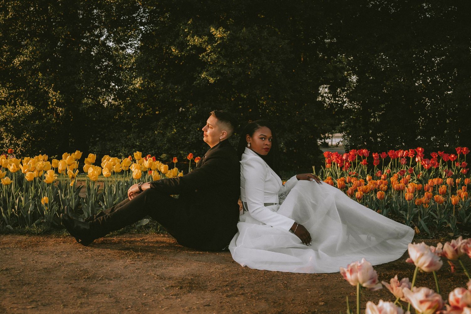 A couple in white outfits sits back to back among colorful tulip gardens.