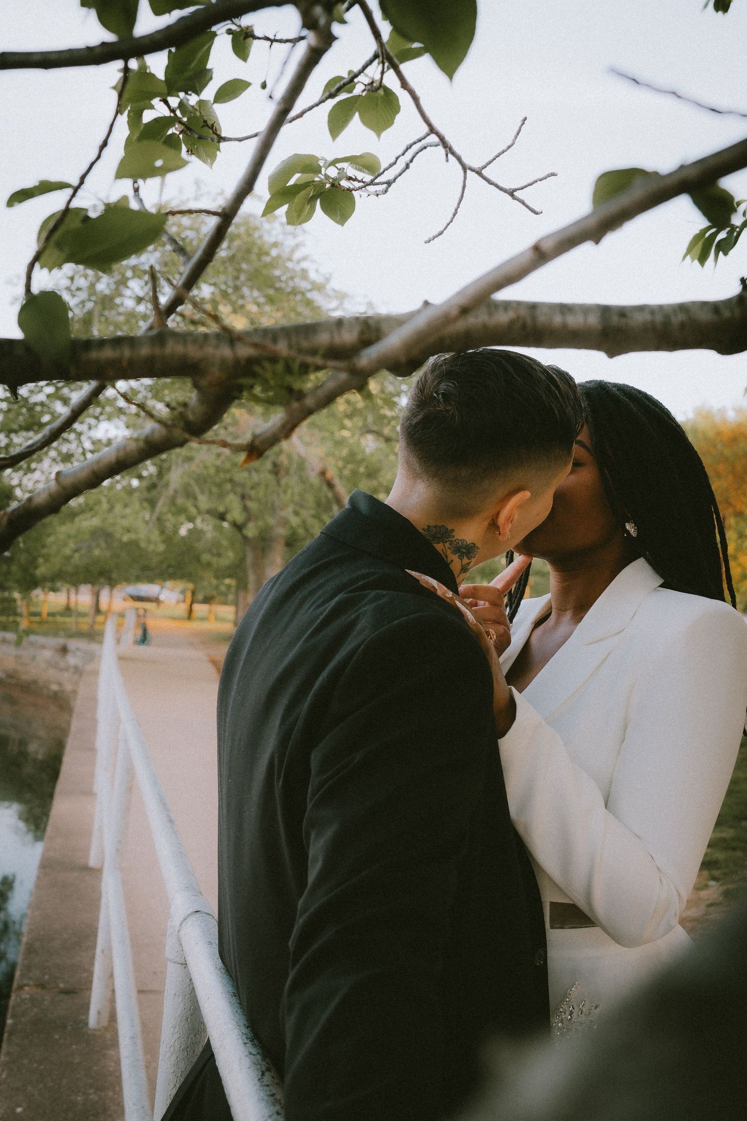 A couple shares an intimate moment under tree branches near a railing.
