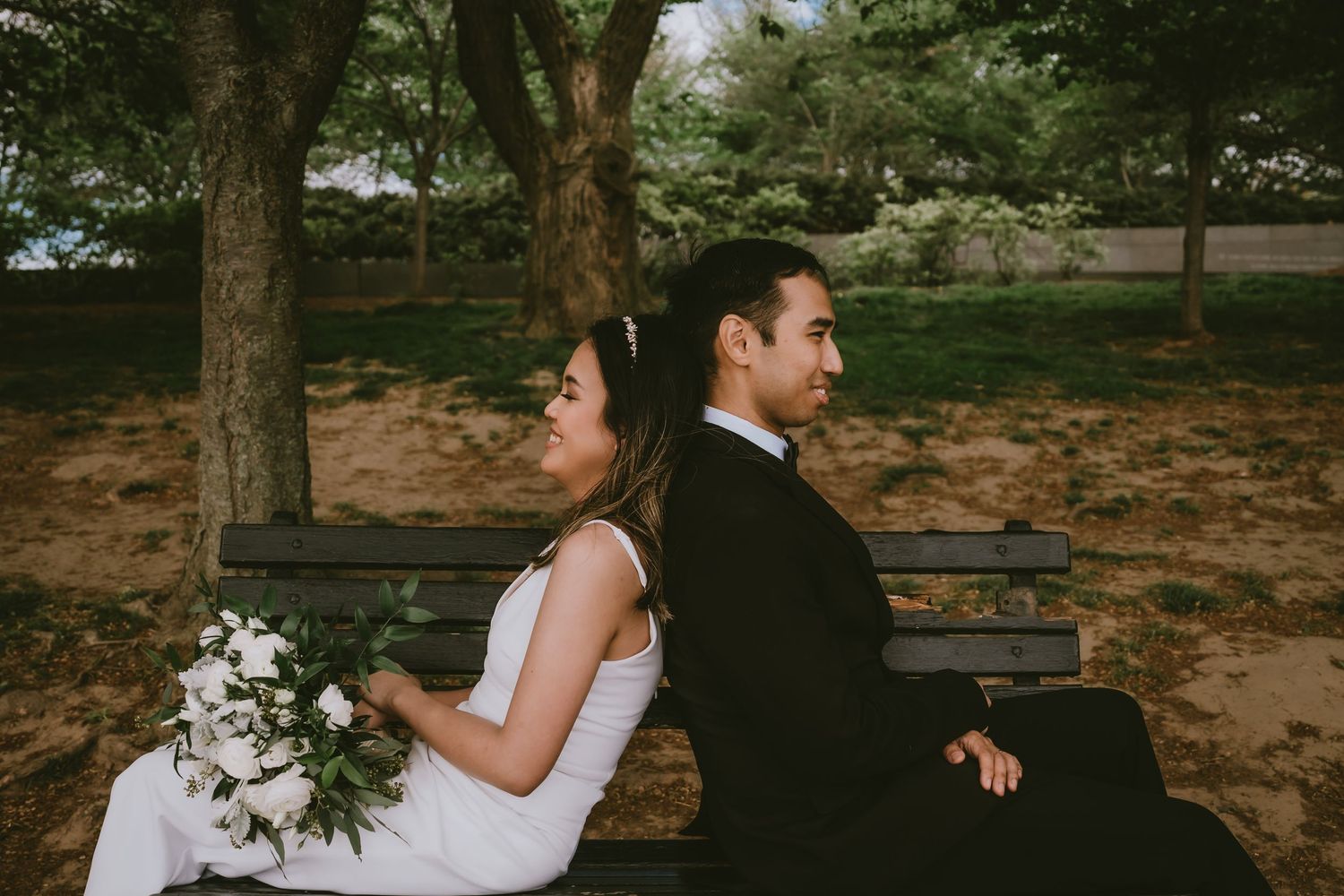 A couple sits back to back on a wooden bench in a peaceful park setting.