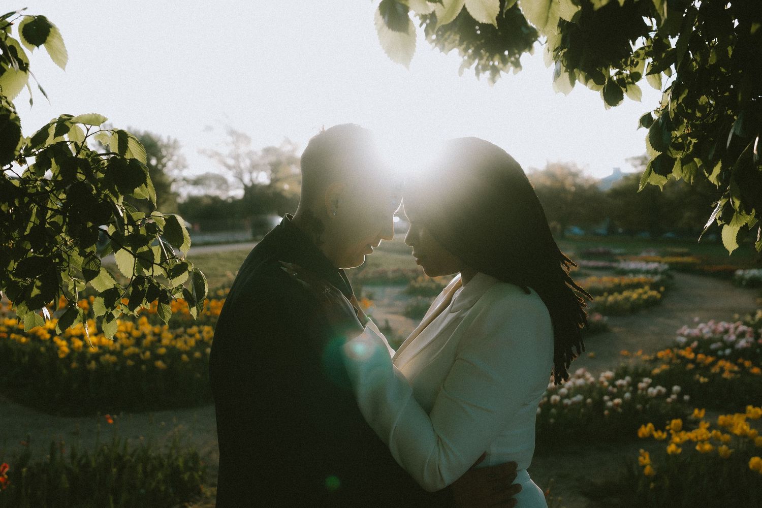 A couple embraces in a garden with sunlight streaming through the trees.