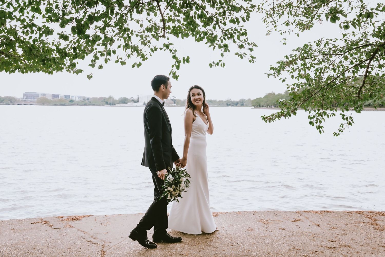 A couple walks along a waterfront path carrying flowers under blooming spring trees.