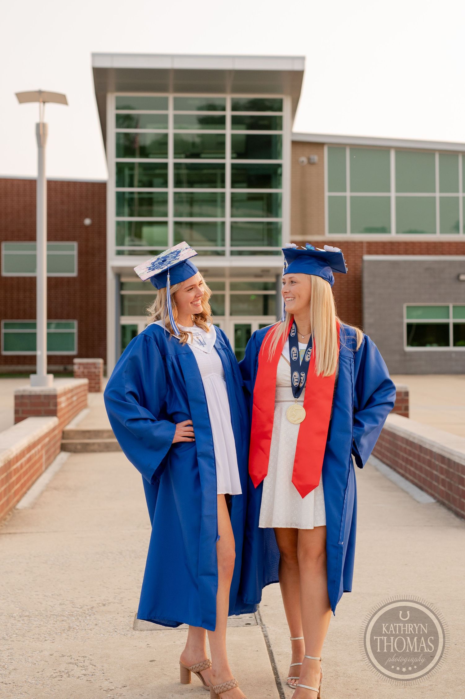 Two graduates in blue caps and gowns sharing a laugh outside their high school.