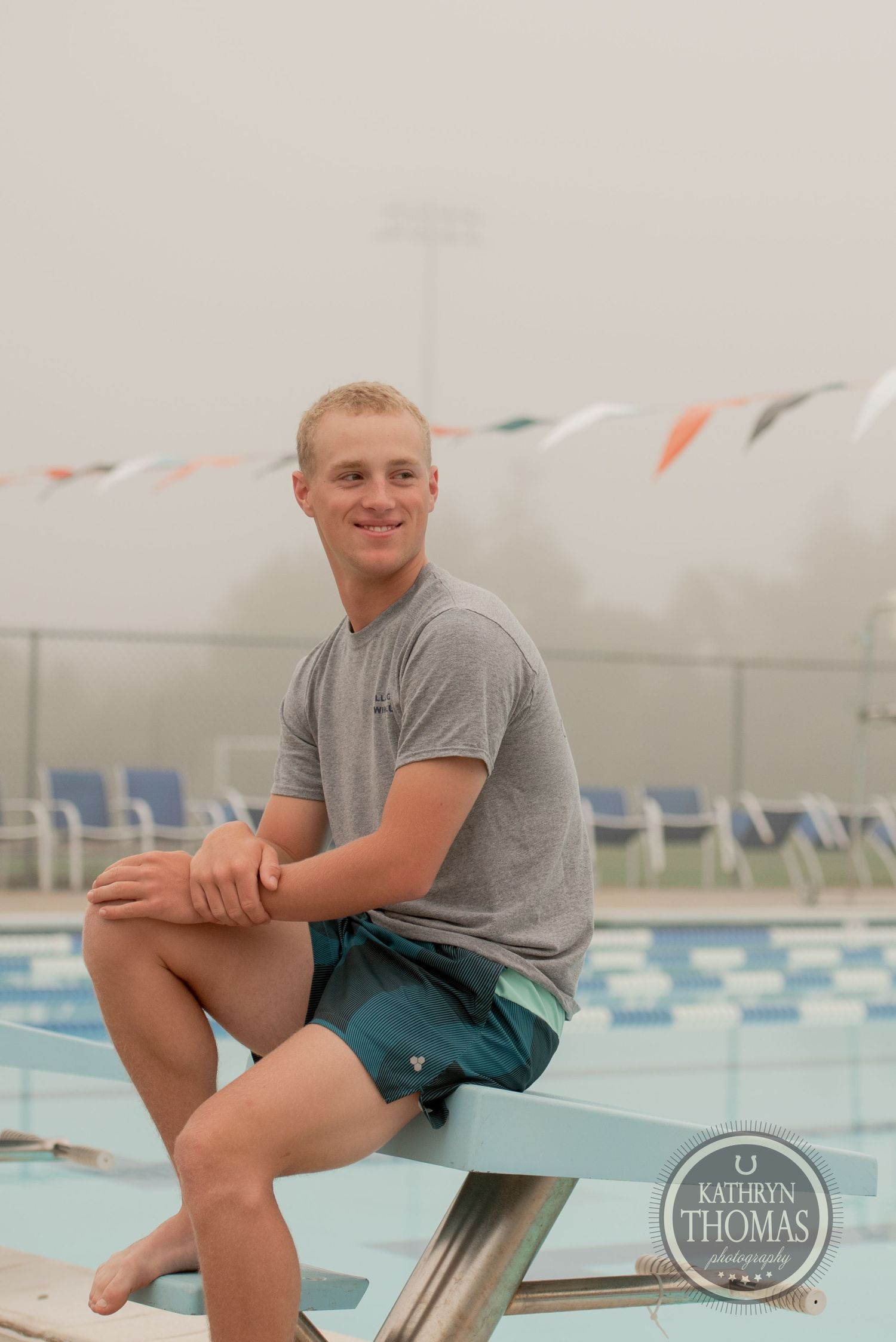 A swimmer in casual attire sits on diving board with pool and colorful flags in background.