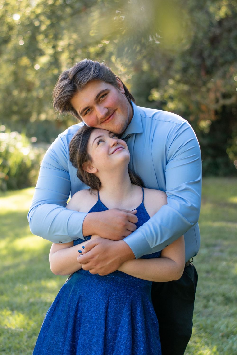 A romantic couple embraces outdoors in a park with sunlit trees in the background.