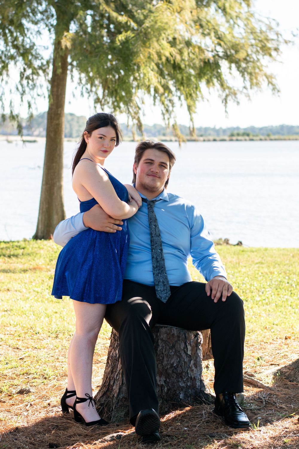 A couple poses together by a tree trunk near a scenic waterfront on a sunny day.