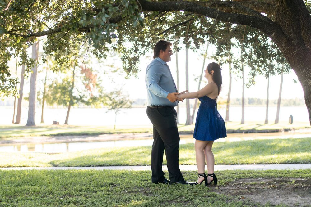 A couple shares a romantic moment under a large tree by a lake during sunset.