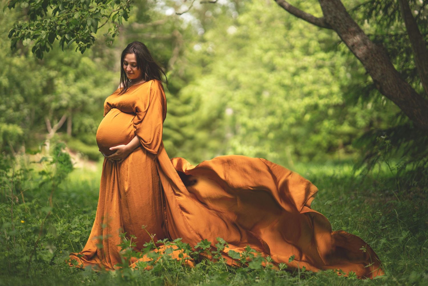 En gravid kvinna i orange silkesklänning där tyget fladdrar i vinden. Hon tittar ner på magen. Hon står i grönskan.