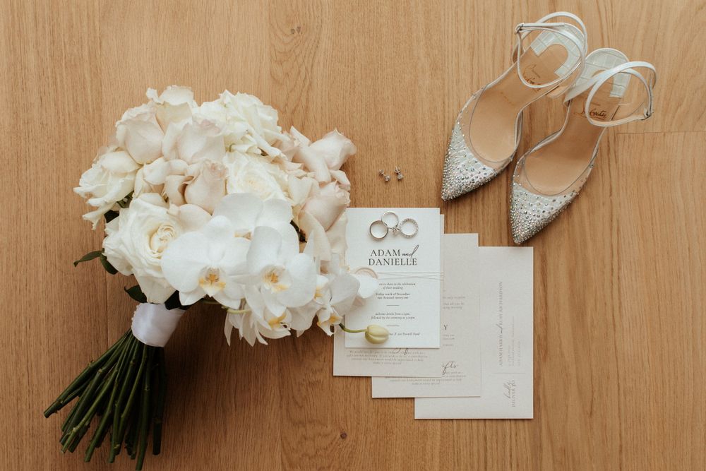 White peony bouquet with sparkly bridal shoes and wedding invitation cards on wooden surface.