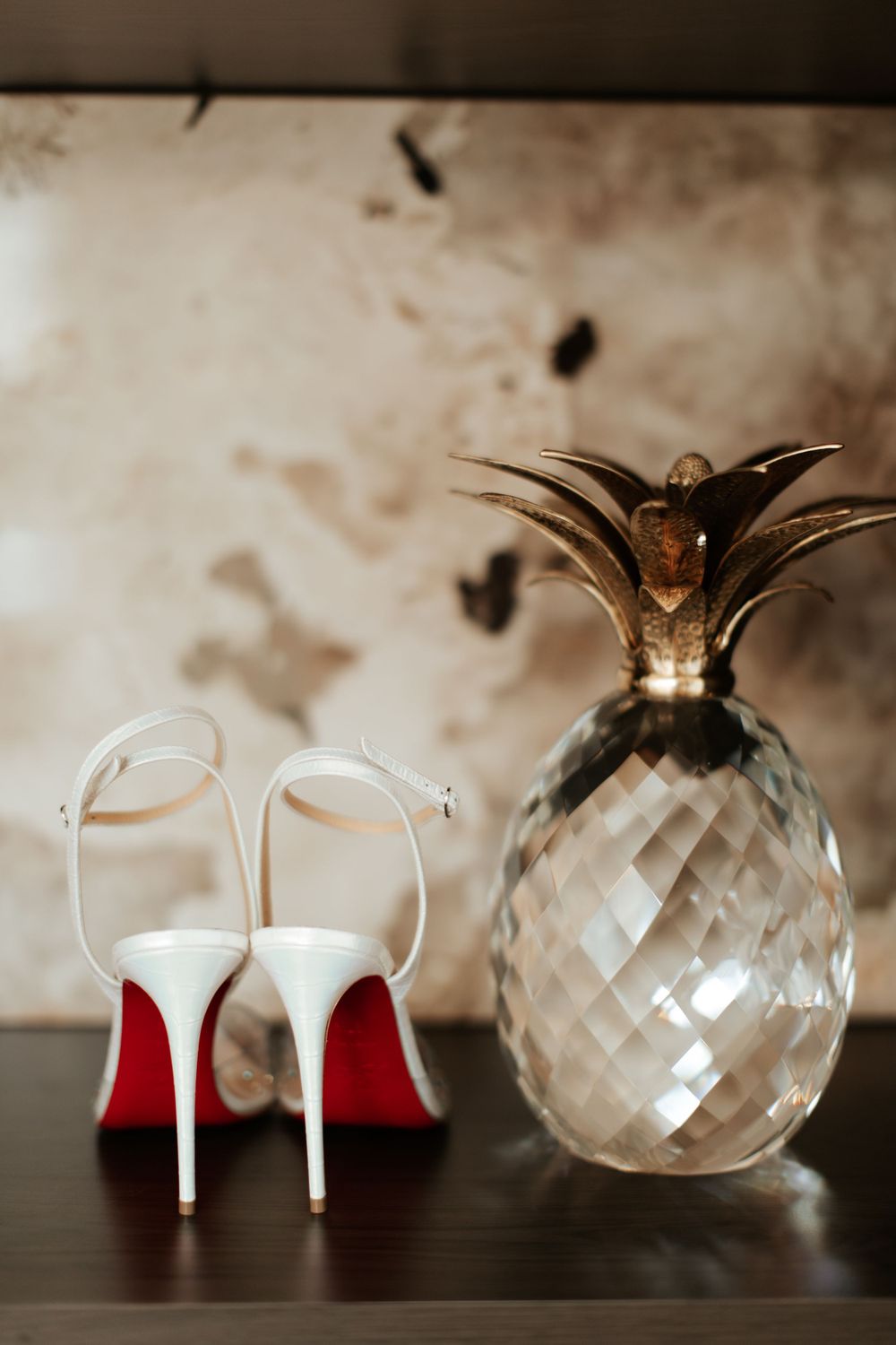 White Christian Louboutin heels next to a decorative crystal pineapple against a dark background.