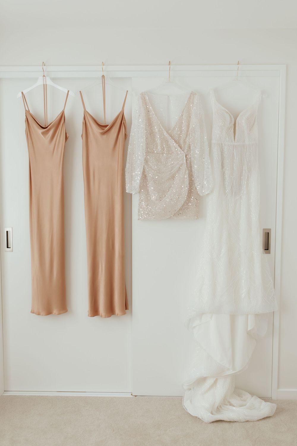 Wedding attire display showing blush bridesmaid dresses and white wedding gown hanging on closet door.
