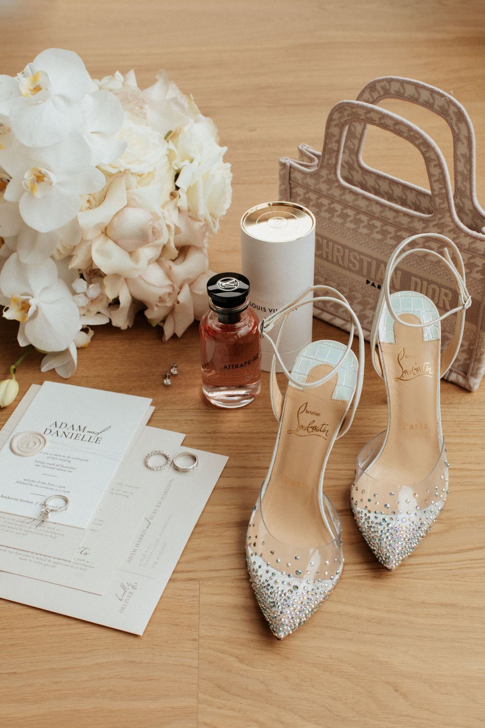 Wedding accessories including crystal heels, white flowers, and perfume arranged on wooden surface.