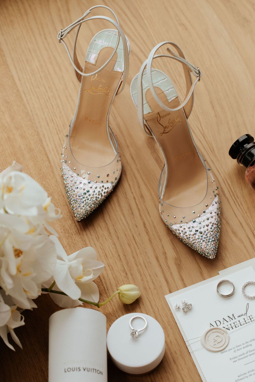 Crystal-embellished bridal shoes with wedding rings and invitation on wooden surface.