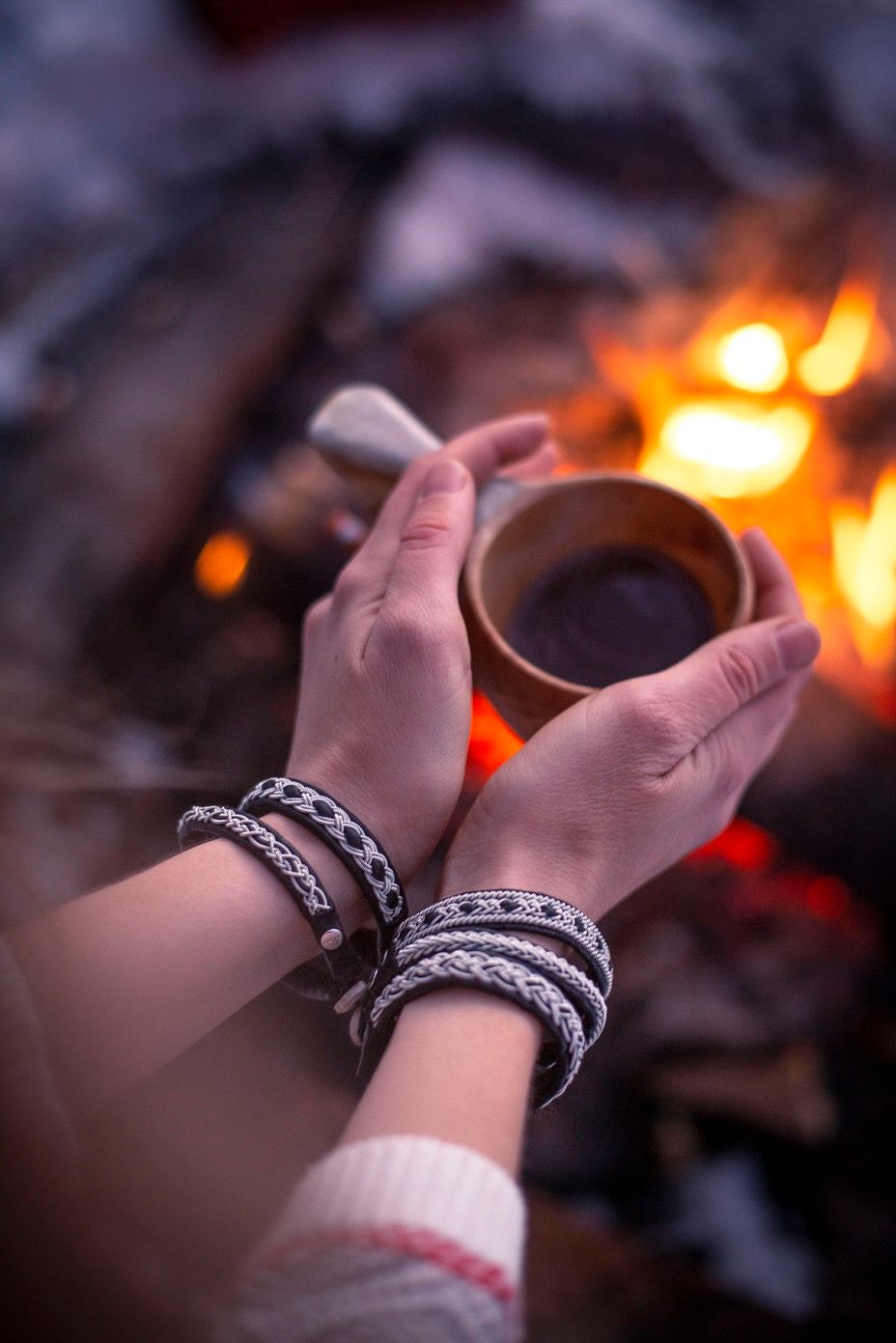 Hands holding a wooden cup of coffee near a glowing campfire with Sami-style bracelets.