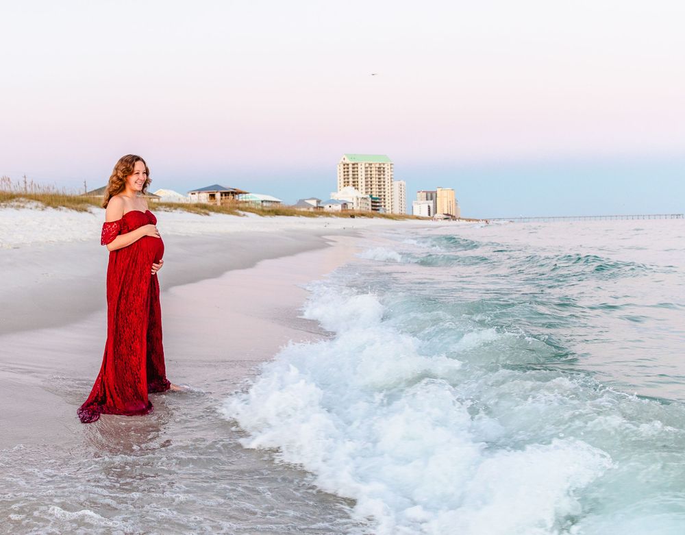 Figure in a flowing red dress stands on a beach at sunset with coastal buildings visible in the distance.