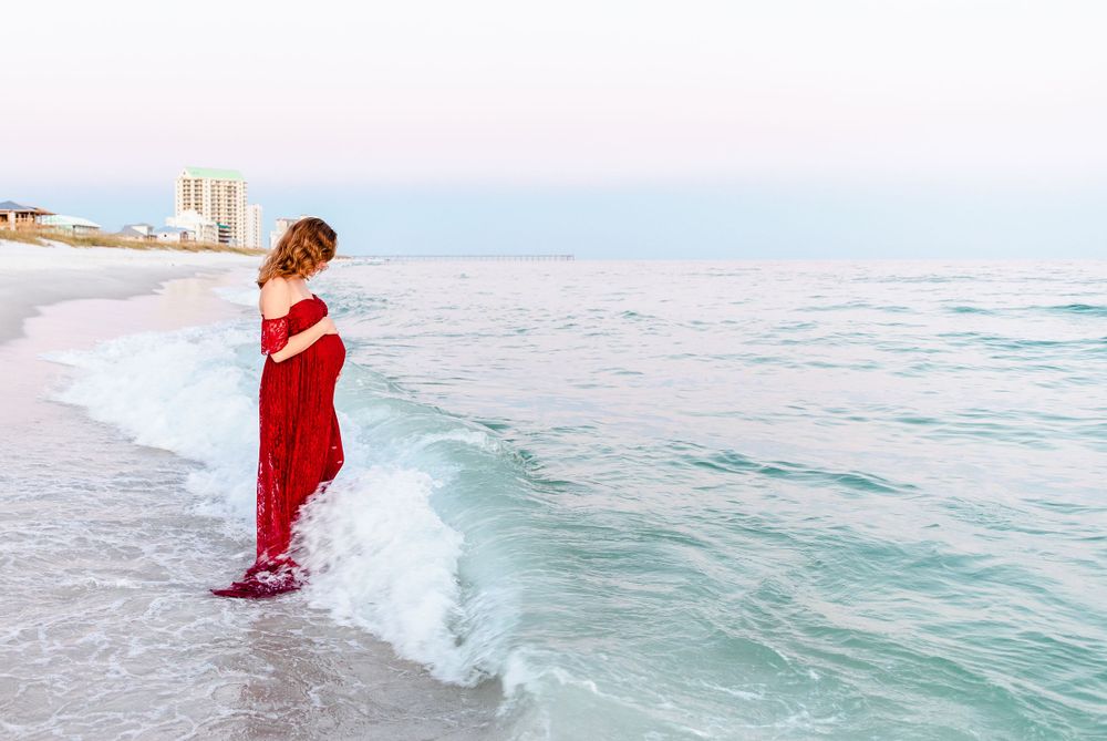Person wearing a red dress stands at the edge of the ocean waves during sunset.