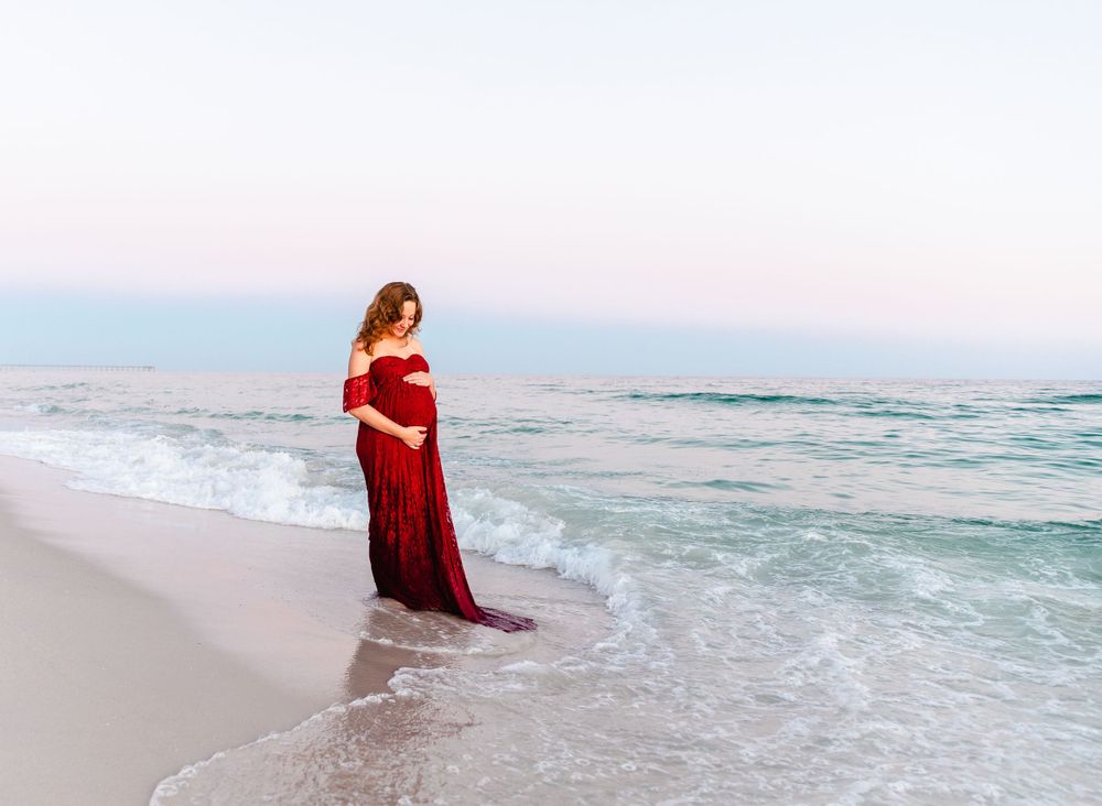 Stunning maternity beach photo with woman in red dress against crashing waves and coastal buildings.