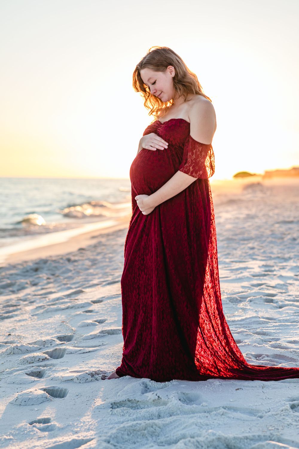Person in red lace dress poses for a maternity photo on a sandy beach at sunset.
