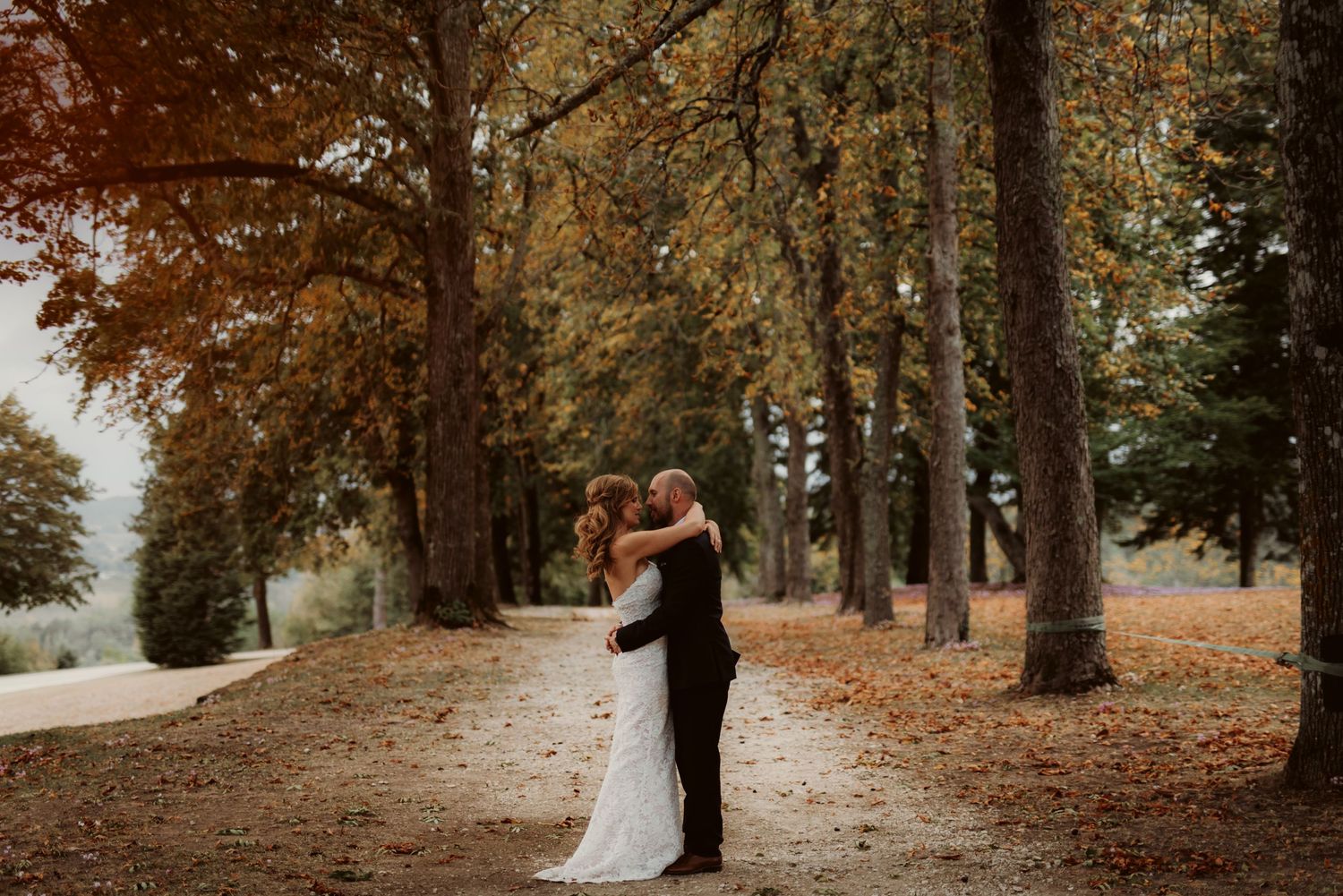 Newlyweds share an intimate moment on a fall-colored path lined with tall trees.