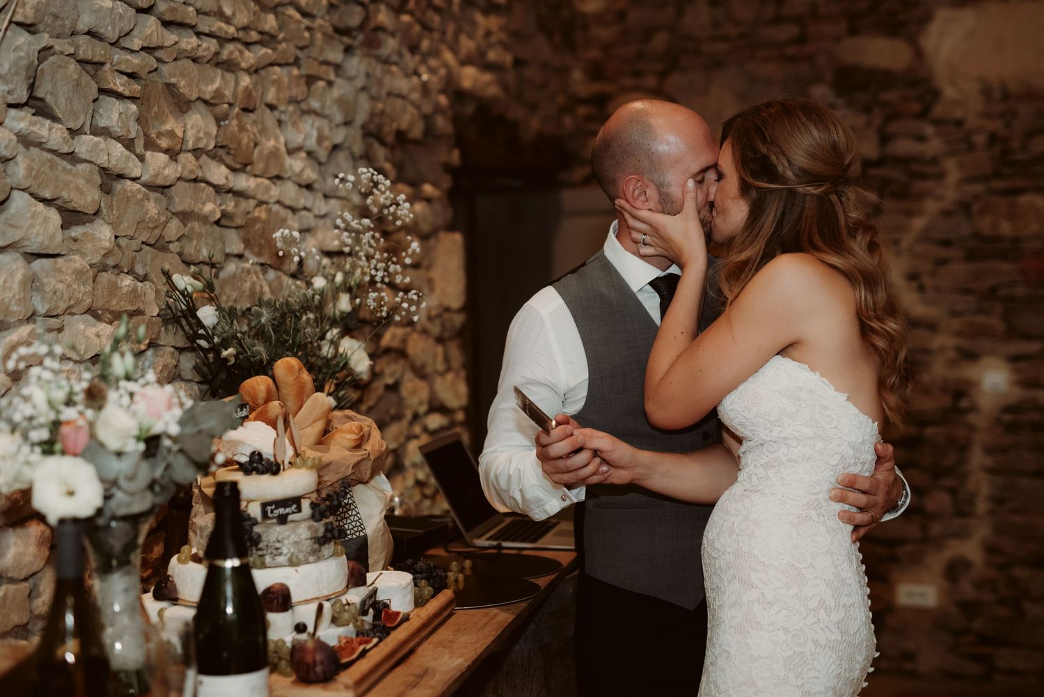 Couple embraces while cutting cake at their wedding reception with floral arrangements and stone walls.