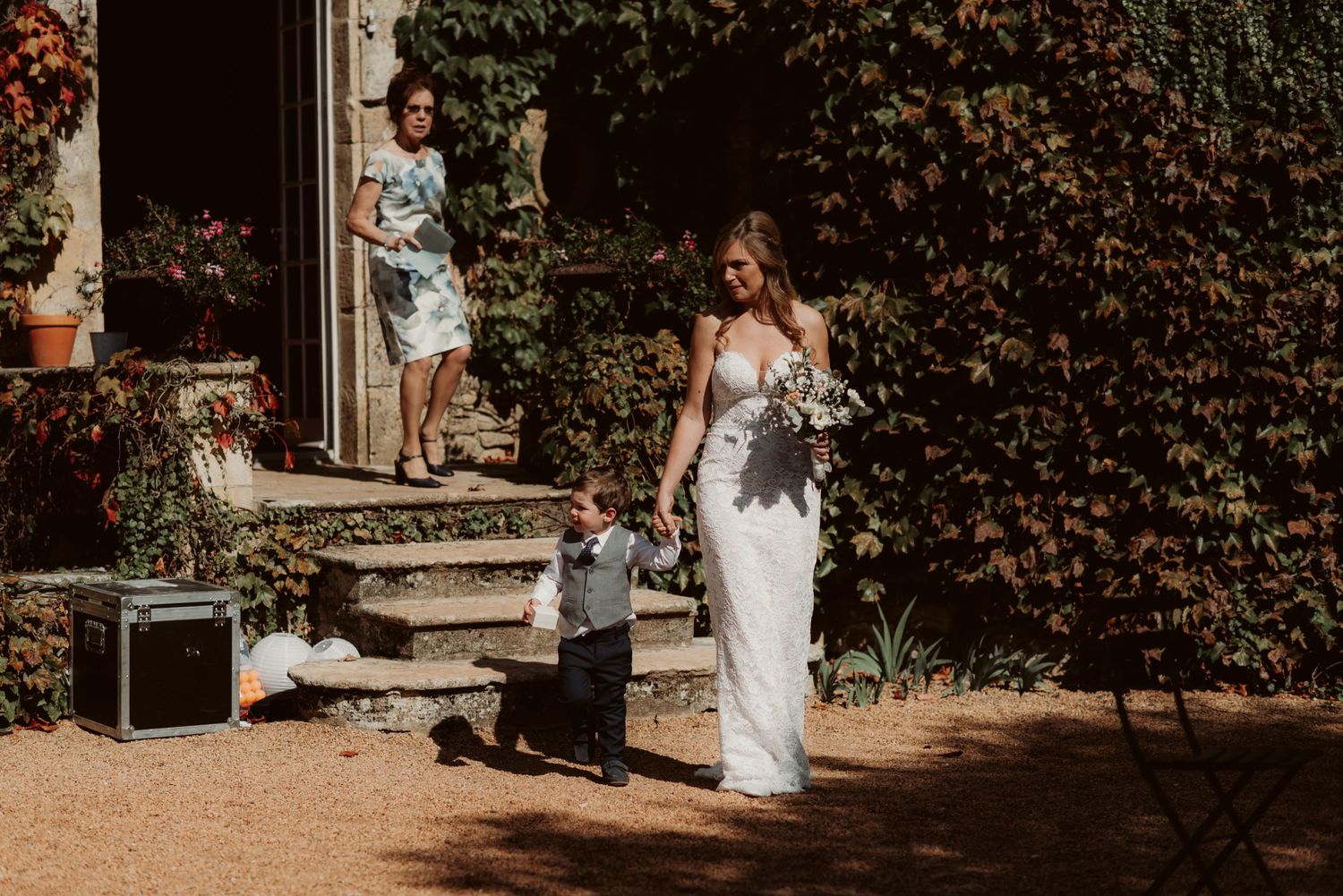 A bride walks along an ivy-covered building in sunlight holding her dress.