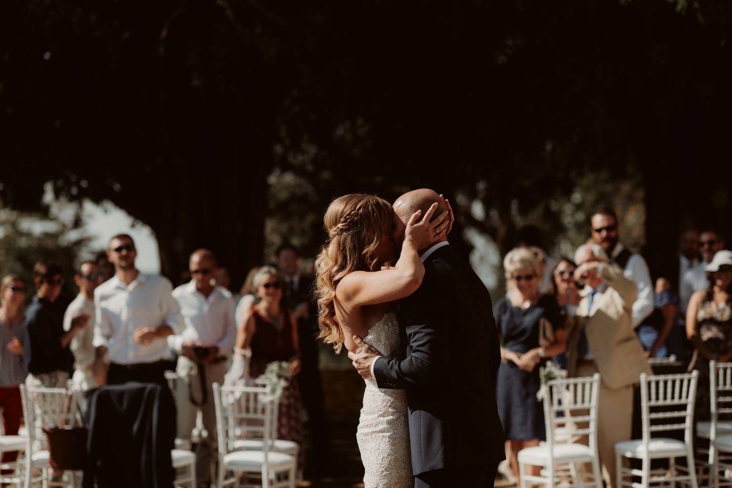 A couple shares their first kiss as newlyweds surrounded by guests in white chairs at an outdoor ceremony.