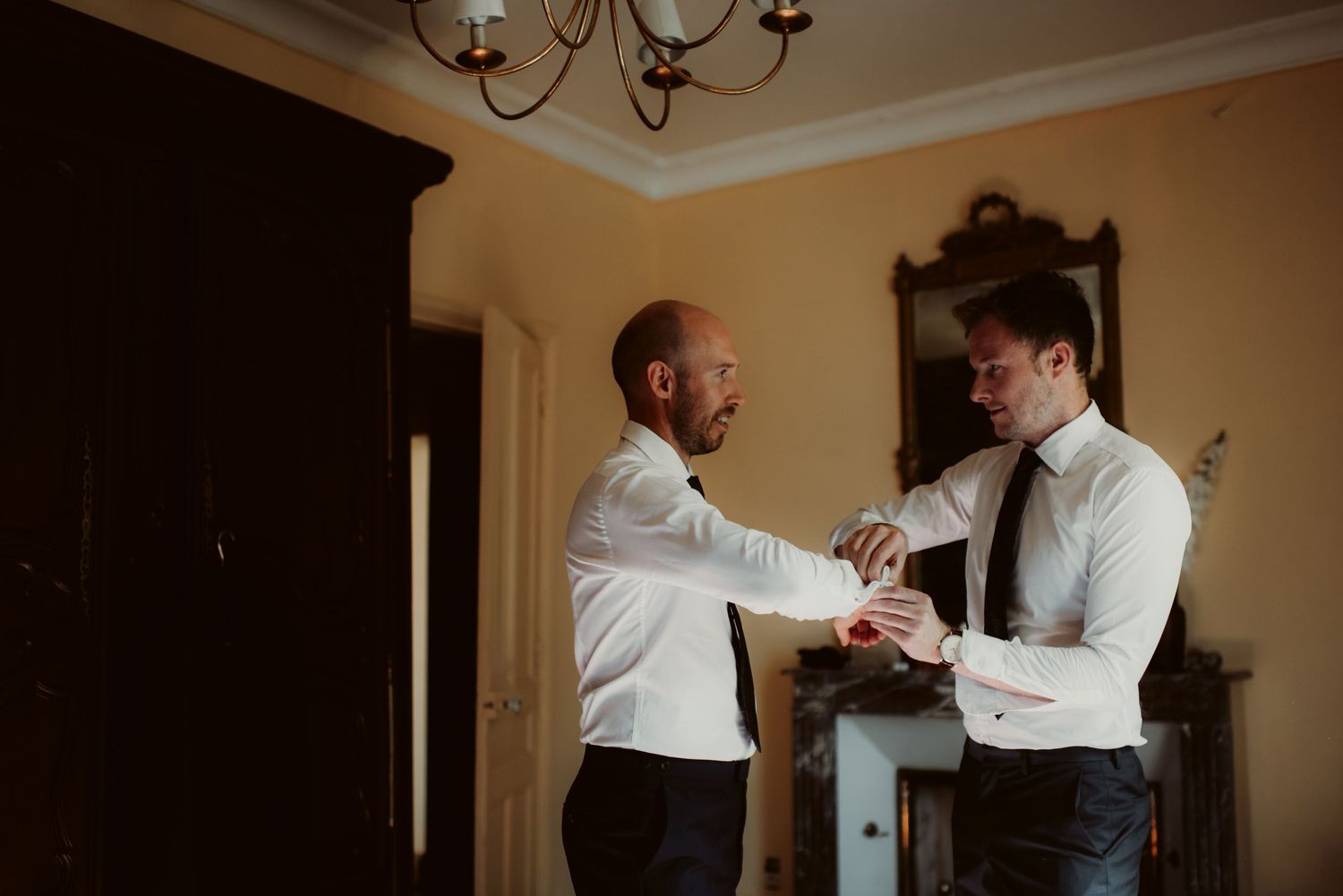Two men in white dress shirts helping each other with formal attire in a warm-lit room.