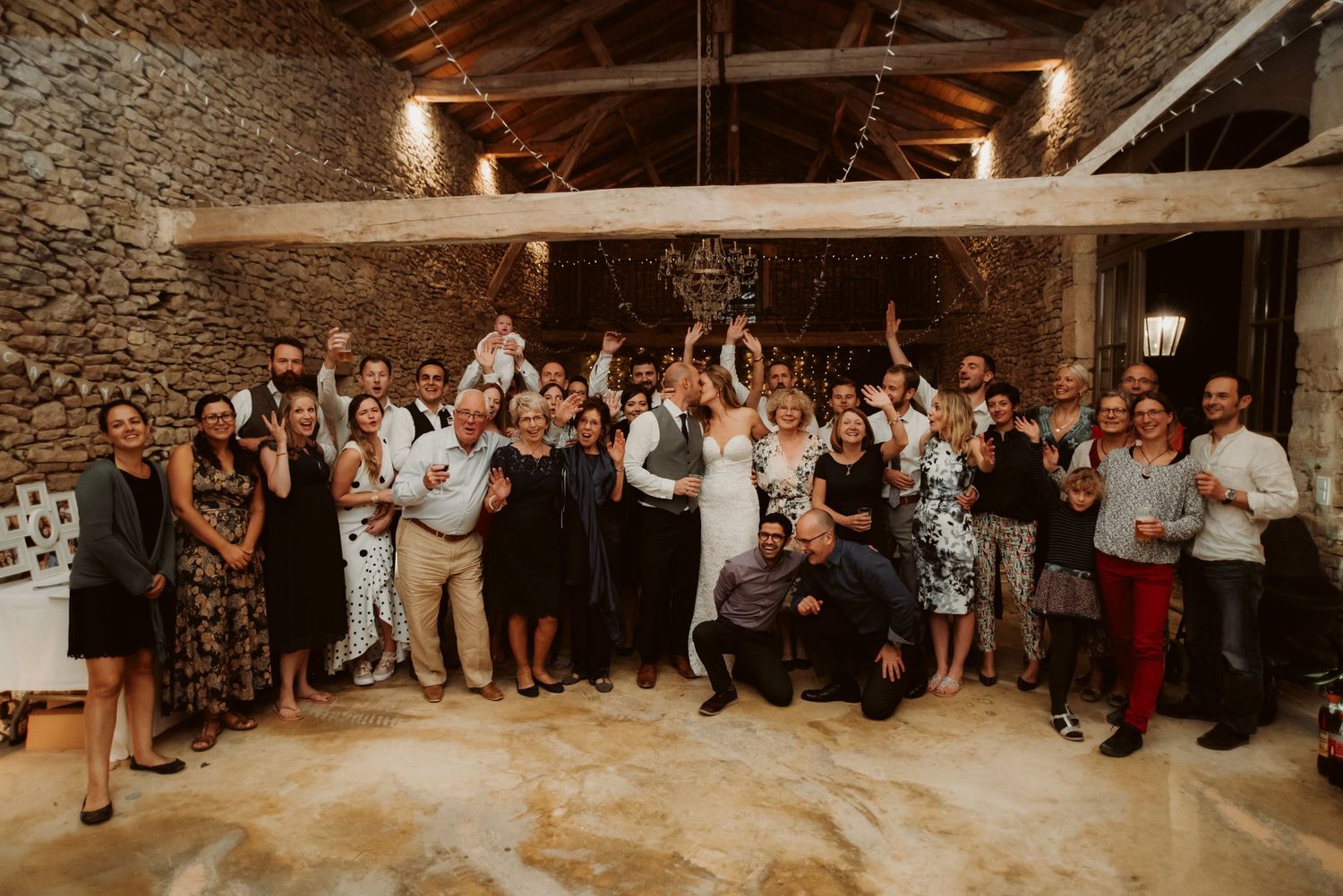 Wedding guests gather for a celebratory group photo in a rustic stone-walled venue with string lights overhead.
