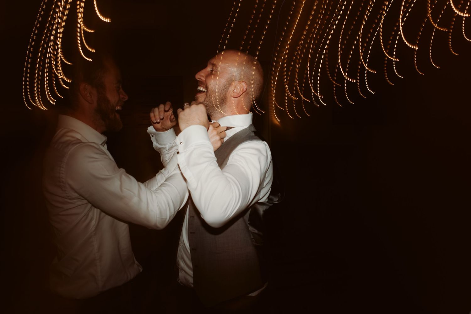Two guests share drinks under warm string lights at an evening reception.