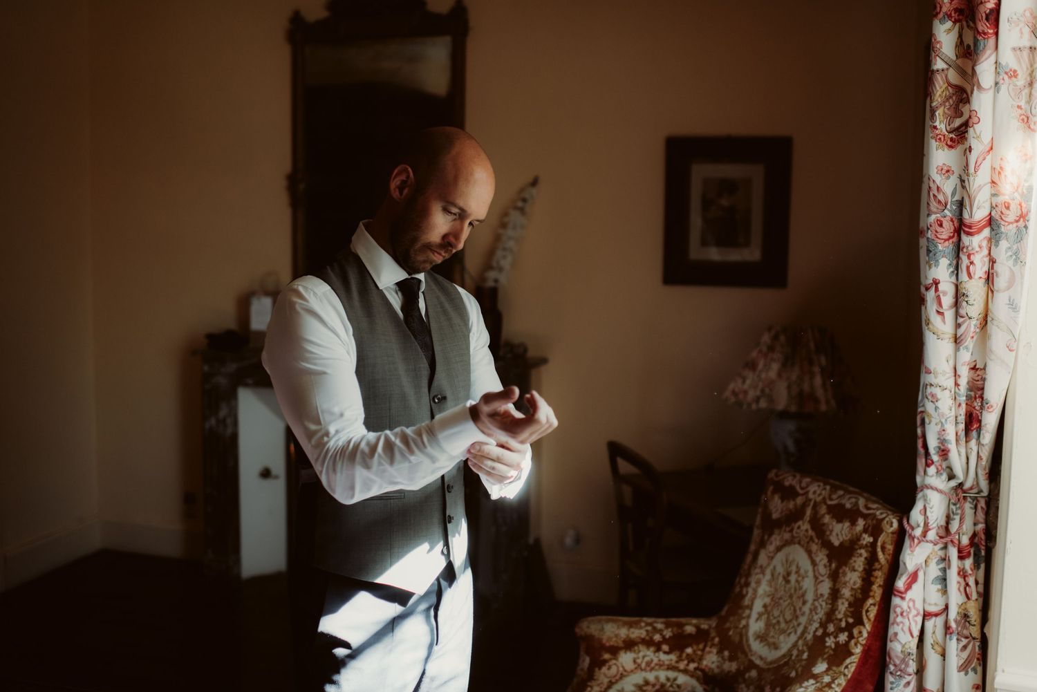 A person in a formal vest checks their phone while standing in a warmly lit room with vintage decor.