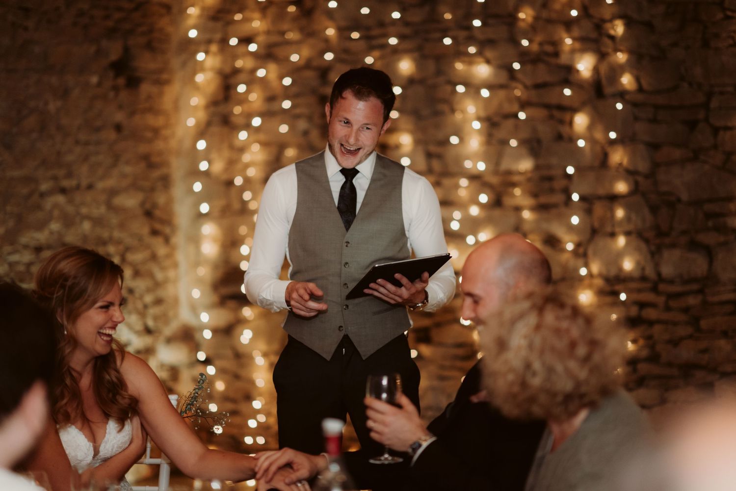 At a wedding reception, a person in a vest reads from a tablet in front of stone walls decorated with string lights.
