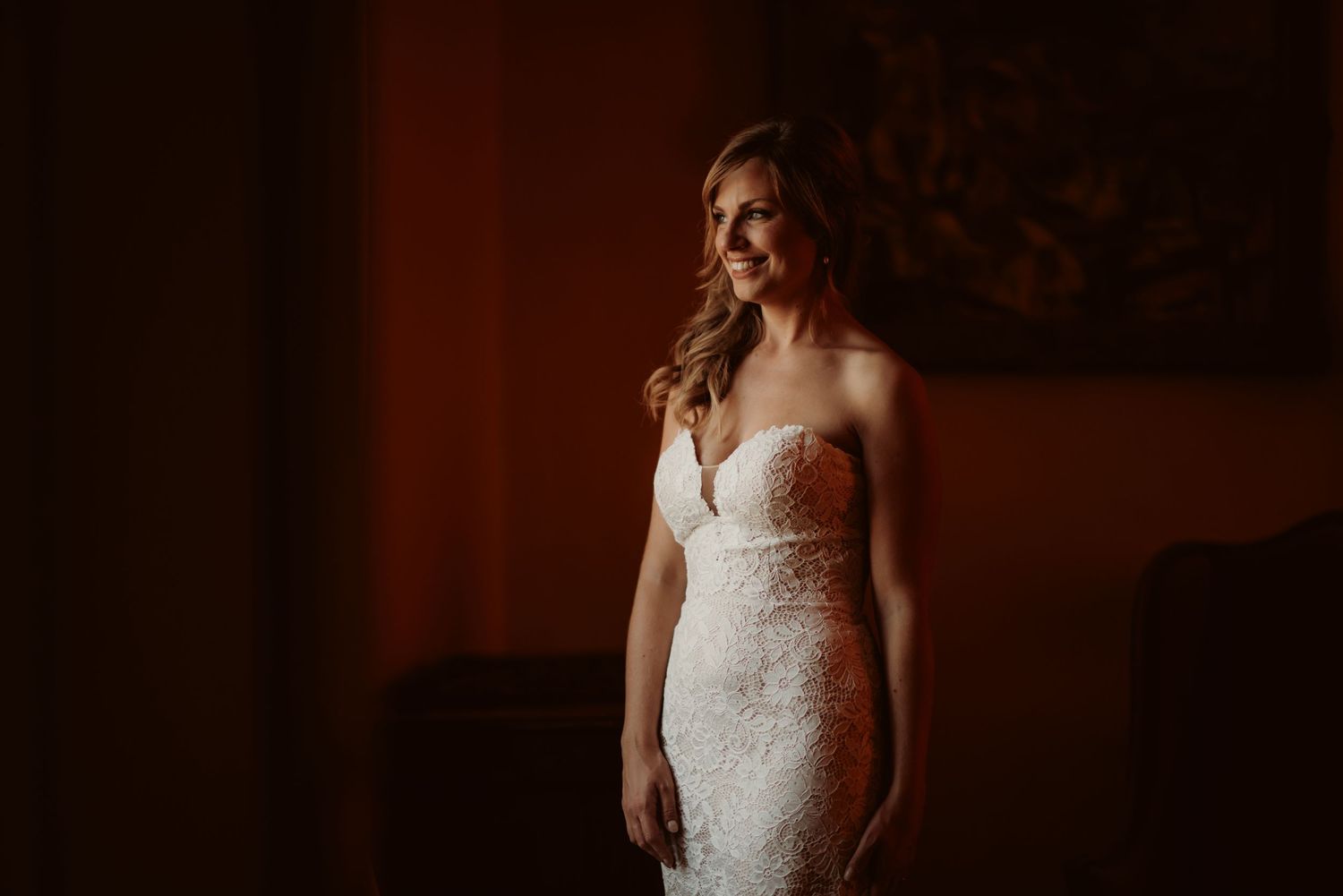 Elegant white lace wedding dress captured in moody lighting against red wall.