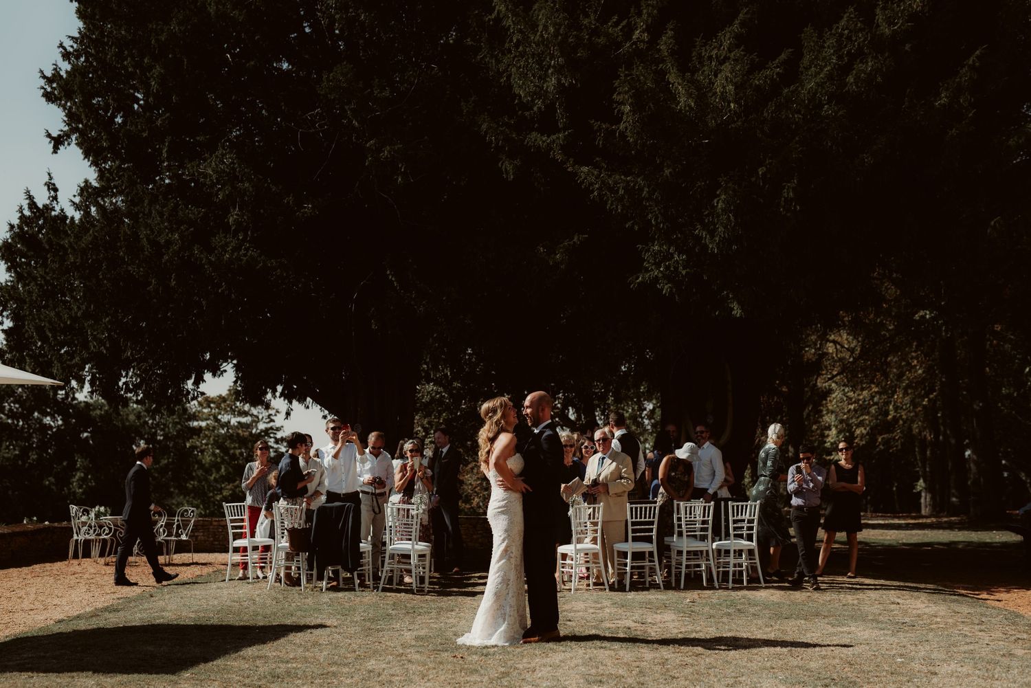 A wedding celebration in a sunny outdoor setting with white chairs and gathered guests.