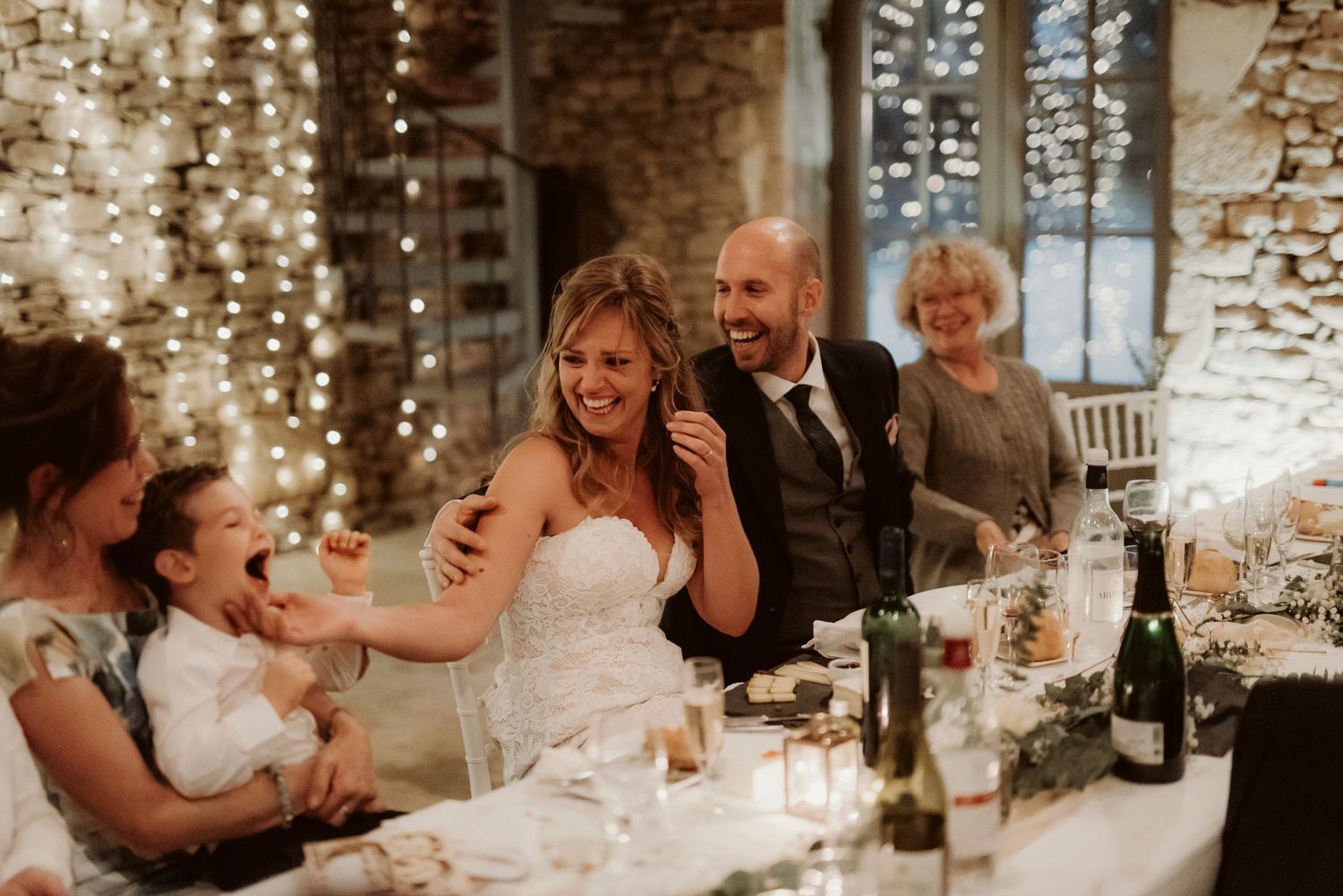 Laughter fills the air during wedding speeches at an intimate stone venue with string light decorations.