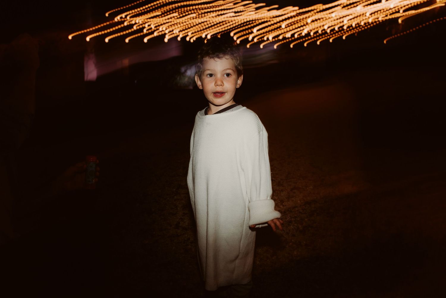 Young guest in white formal attire stands beneath decorative string lights.