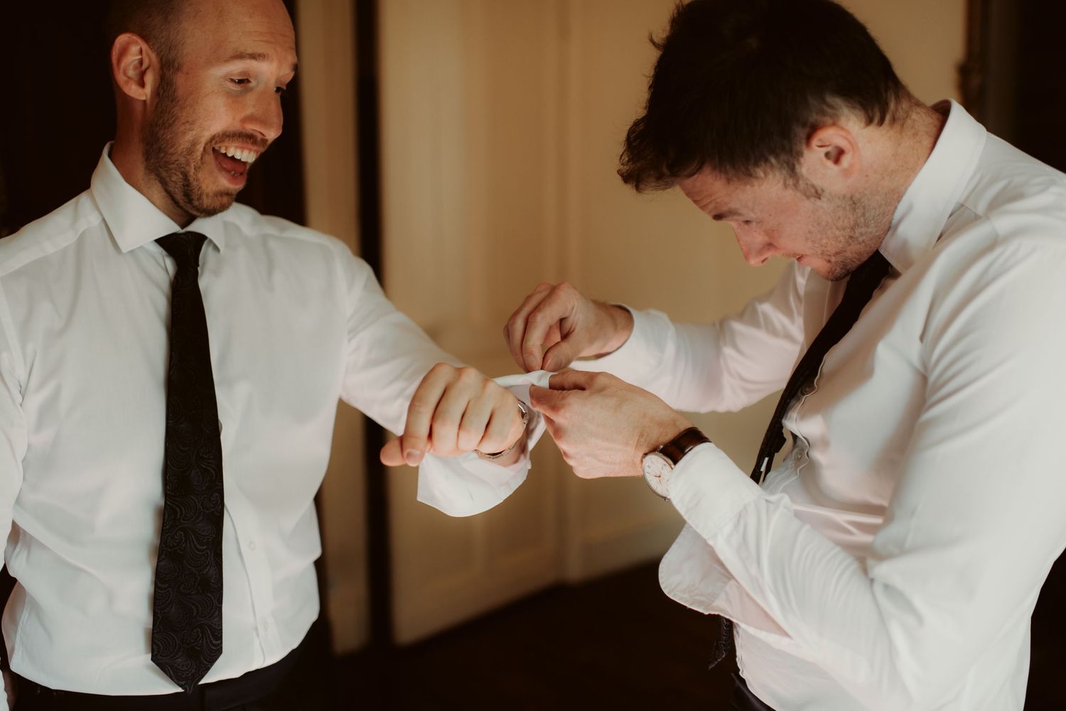 Two men in formal wear helping each other with cufflinks during preparation.