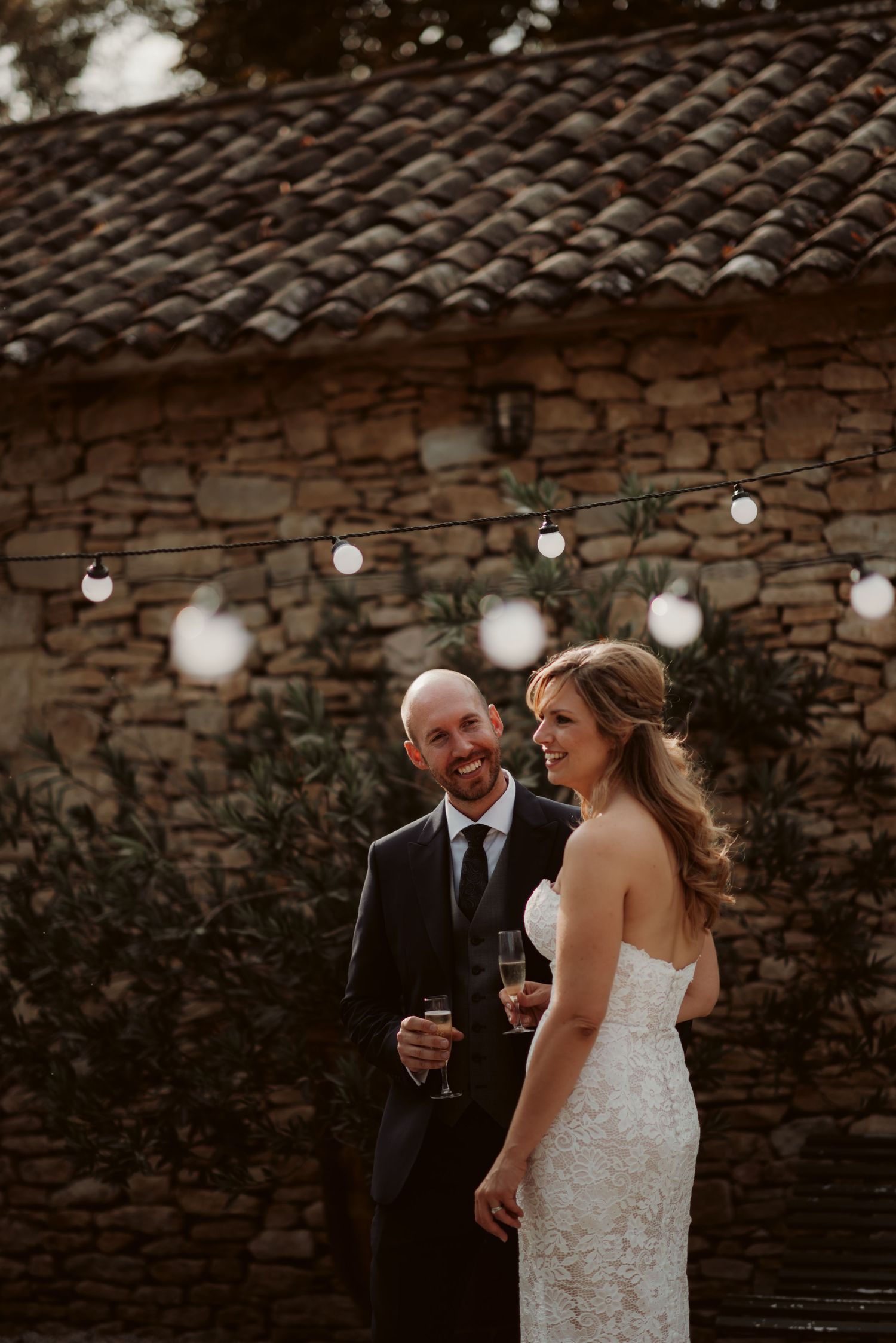 A romantic moment captured at dusk with string lights illuminating a rustic stone wall.