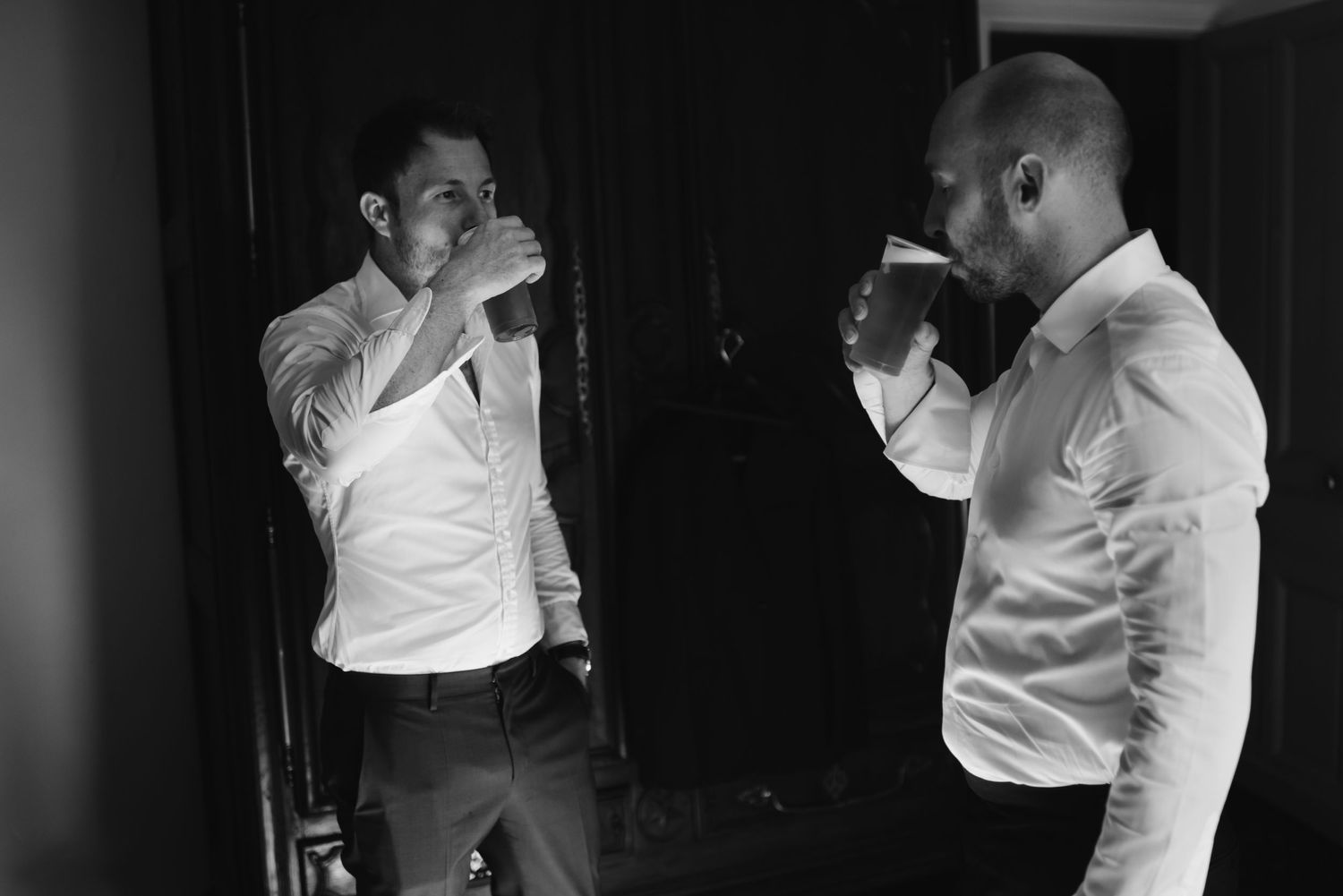 Two men in white dress shirts laugh while adjusting their formal attire in a dark room.