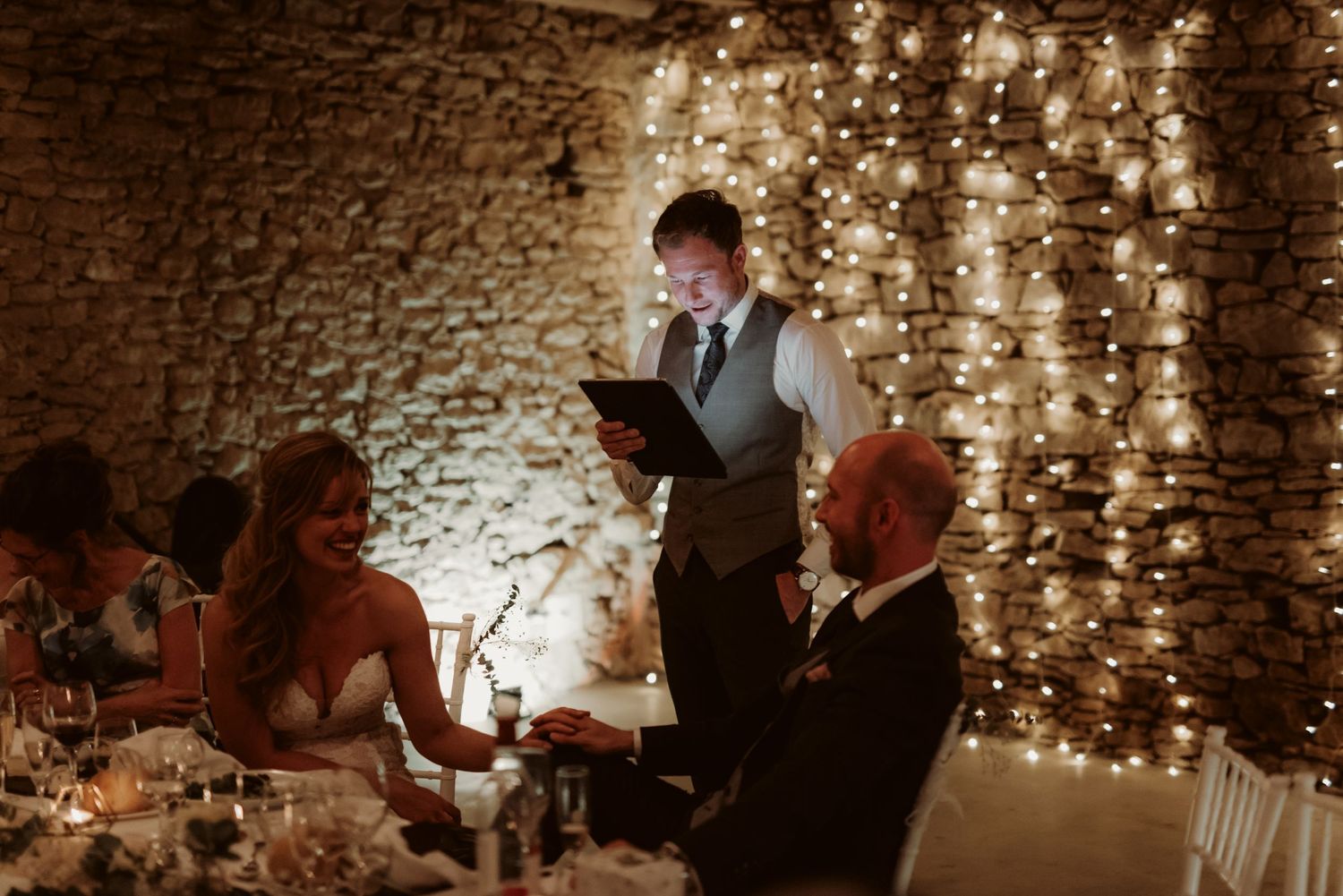 A warm-lit wedding reception scene with string lights against stone walls during dinner service.