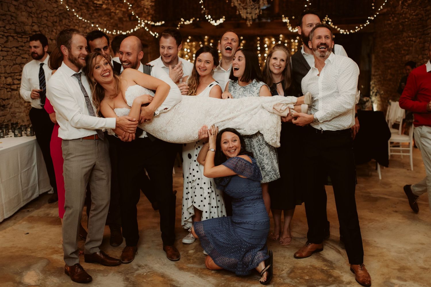 Wedding party lifts and carries celebrants during a fun reception moment in a rustic stone venue.