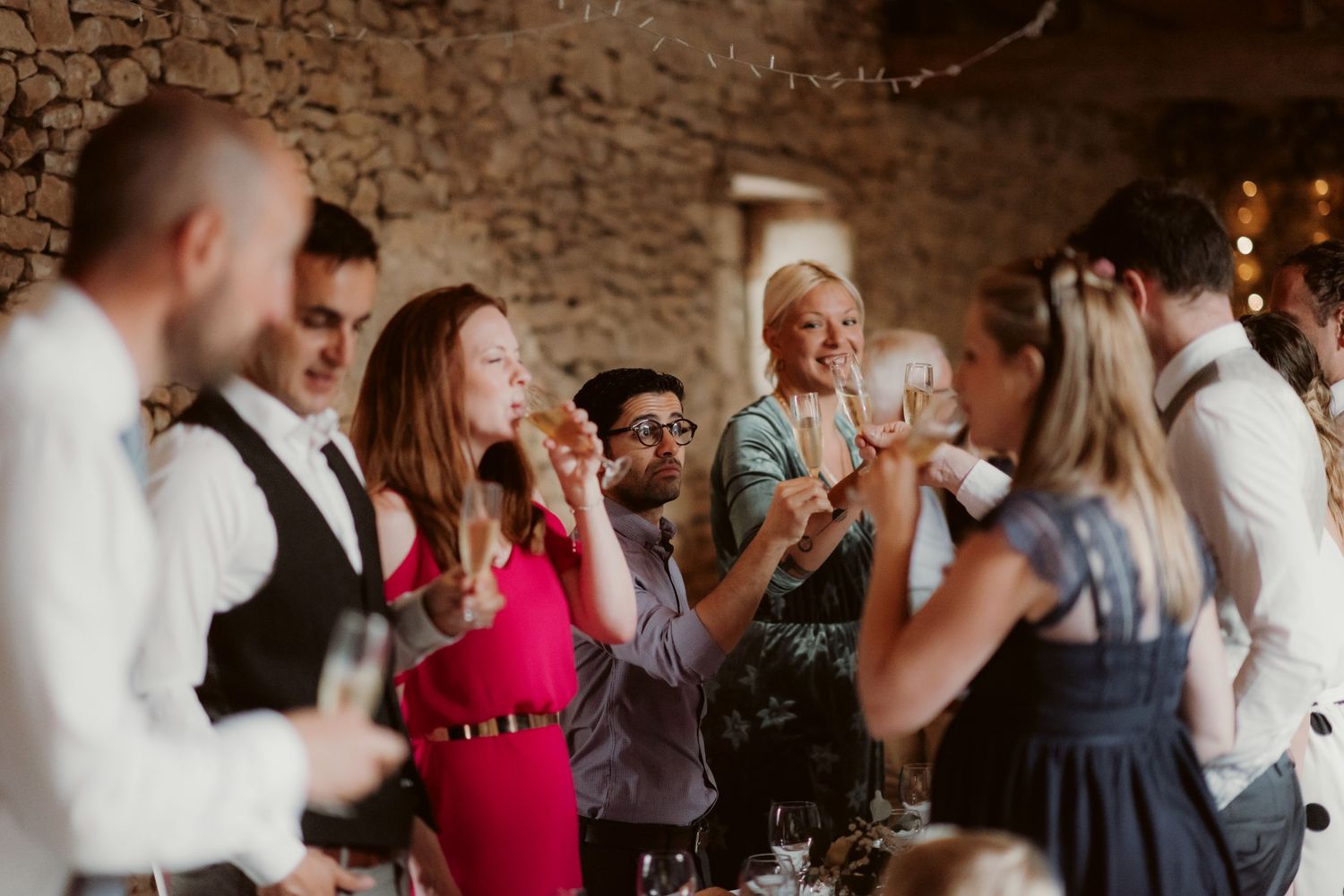 Wedding guests laugh and celebrate together in a rustic stone-walled venue with twinkling lights.