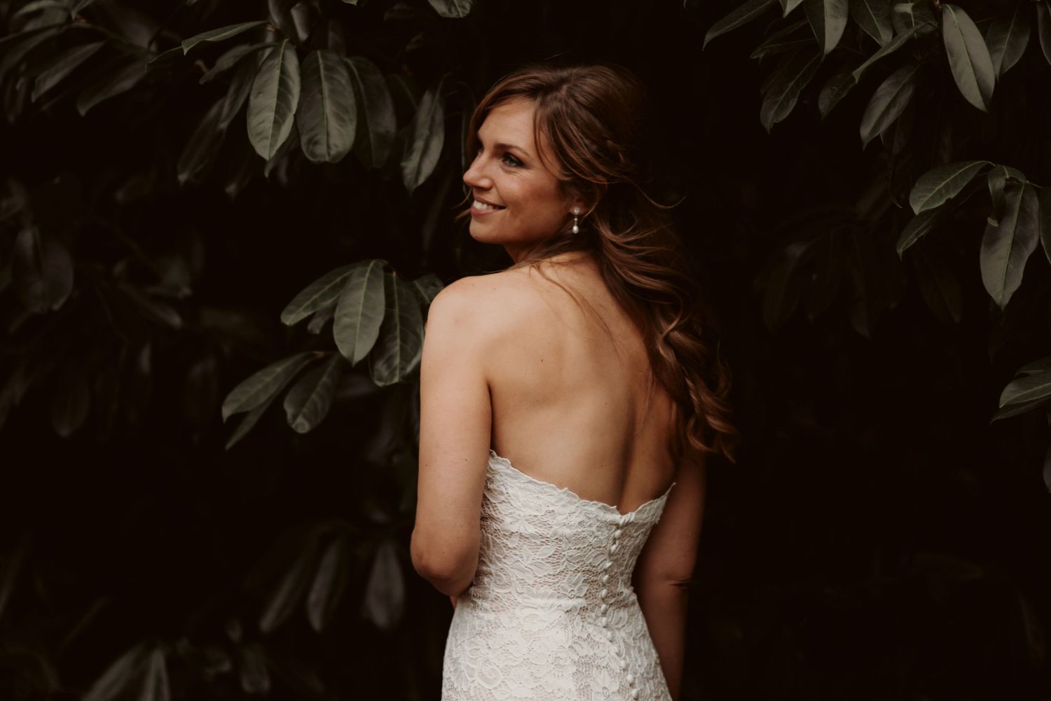 Elegant bride in strapless lace gown poses against dark green foliage backdrop.
